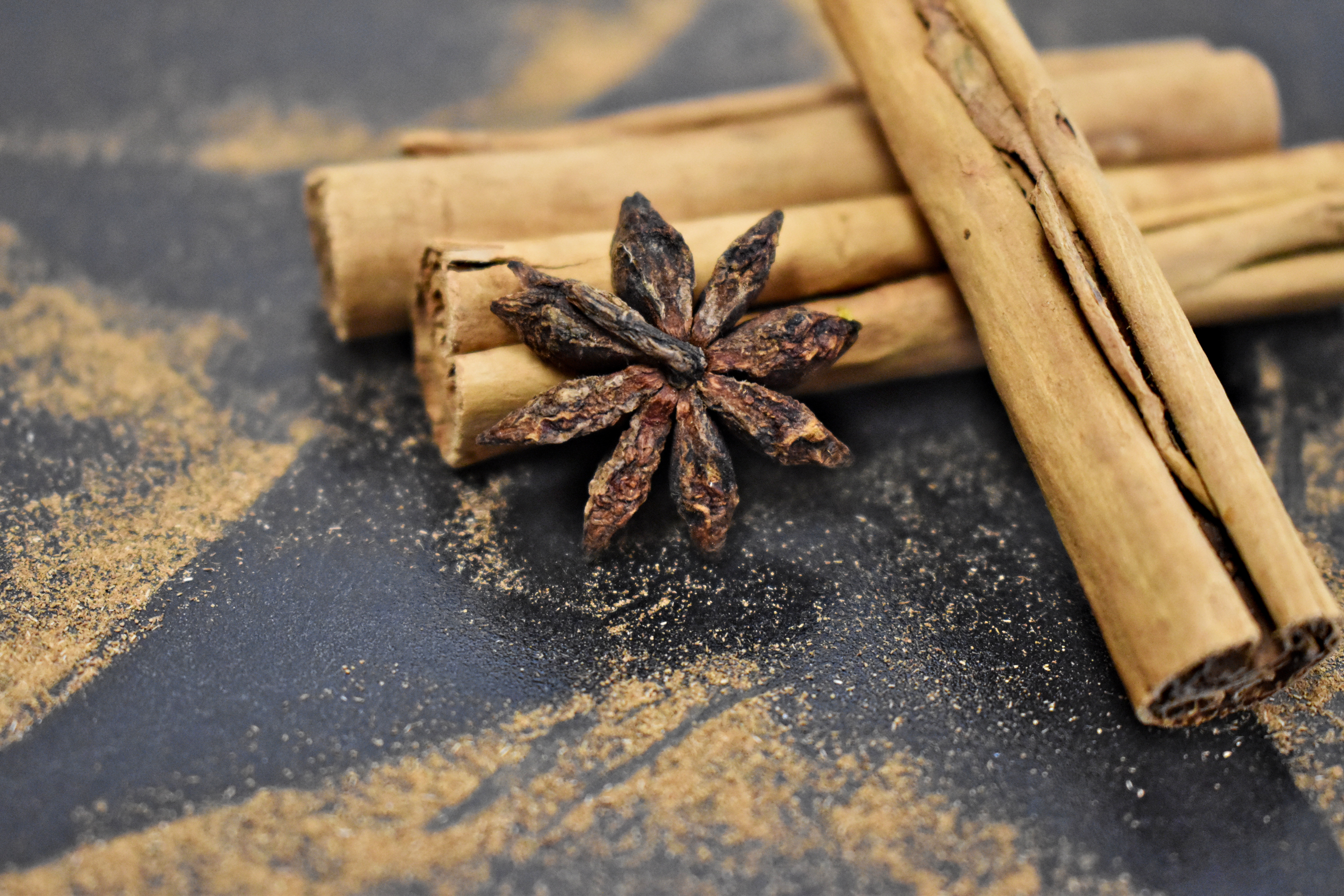Auf diesem Bild sind drei gestapelte Zimtrollen und eine getrocknete Sternanis zu sehen, welche auf einer schwarzen Steinplatte, umgeben von einer Sternenform aus Zimtpulver, liegen.