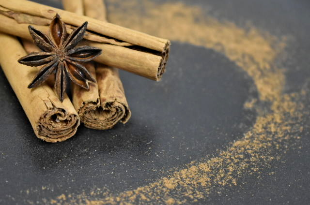 Auf diesem Bild sind drei gestapelte Zimtrollen und eine getrocknete Sternanis zu sehen, welche auf einer schwarzen Steinplatte, umgeben von einer Herzform aus Zimtpulver, liegen.